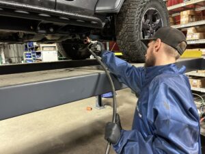 A TNT Customs technician in protective gear sprays undercoating onto the underside of a lifted Jeep inside a workshop, ensuring rust and corrosion protection.