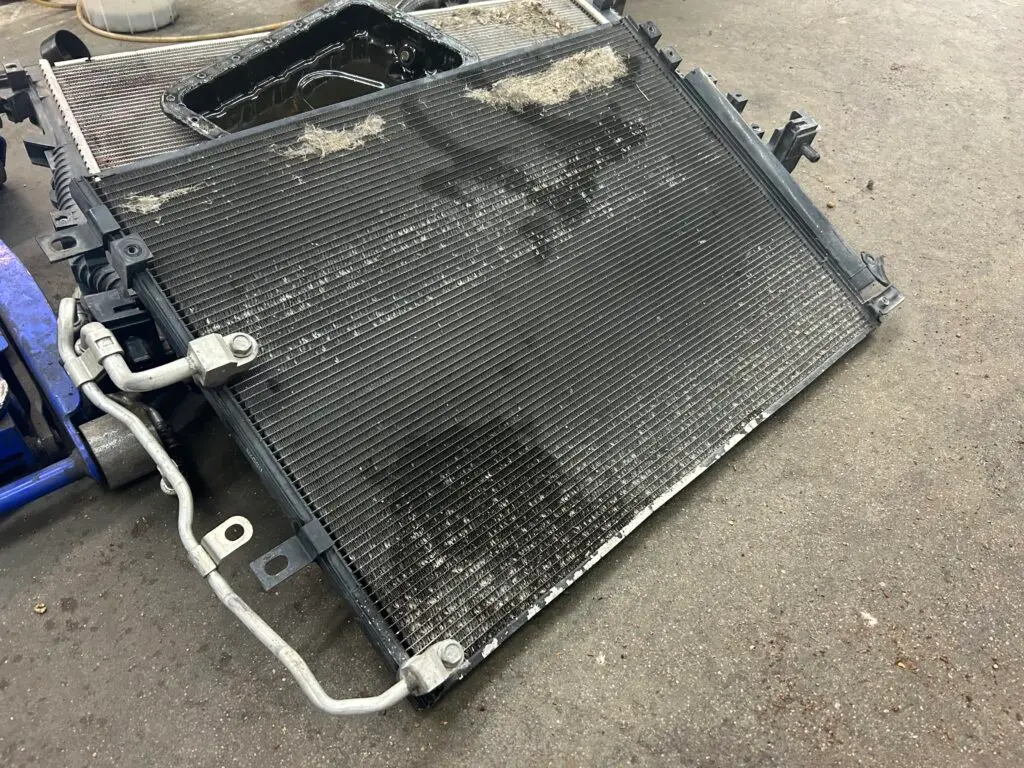 Used, dirty car radiator with visible debris and coolant stains on a shop floor, removed for inspection or replacement.