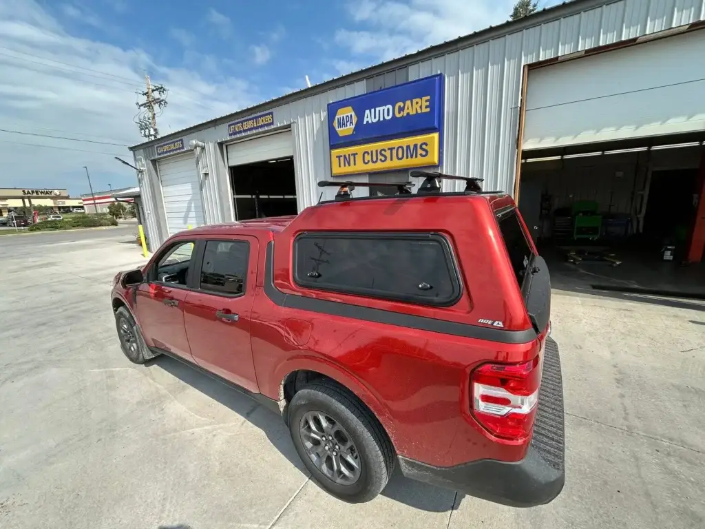 Red truck parked outside TNT Customs Auto Repair shop featuring a newly installed ARE topper with roof rack.